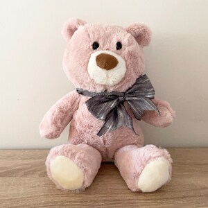 May include: A plush pink teddy bear with a grey and silver bow. The bear has a brown nose and black eyes.