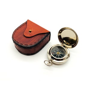 May include: A silver compass with a black face and a leather carrying case. The compass has a needle that points north and is marked with the cardinal directions. The case is brown and has a flap that closes with a button.