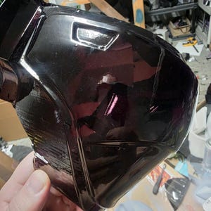 May include: A close-up of a black, glossy, angular object, possibly a piece of armor or a helmet component. The surface reflects light, highlighting its shape and design. The object is held in a person's hand.