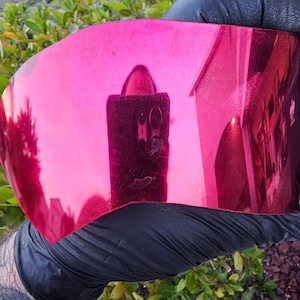 May include: A close-up of a pink, reflective, curved piece of plastic held in a gloved hand. The plastic is reflecting a blurry image of a building and a person.