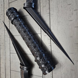 May include: Two black, intricately designed throwing knives with a decorative sheath. The knives have a pointed tip and a handle with a decorative design. The sheath is cylindrical and has a similar decorative design.