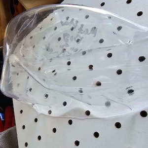 May include: A clear plastic face shield with the words "Matt's Big" written on it. The shield is resting on a white surface with evenly spaced black dots. Other objects are visible in the background.