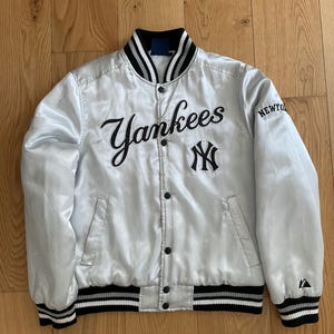 Può includere: Giacca da baseball dei Yankees in raso argentato con finiture a righe bianche e nere su colletto, polsini e orlo. La parola "Yankees" è ricamata in nero sul petto, con il logo della squadra sottostante. La parola "NEW YORK" è sulla manica destra.
