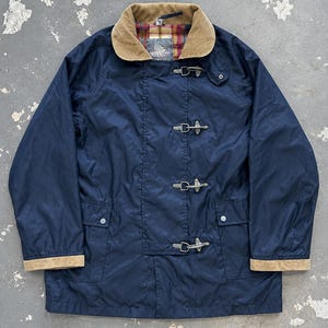 Può includere: Una giacca blu navy con colletto in velluto a coste color marrone chiaro e polsini abbinati. La chiusura frontale è composta da quattro fermagli a levetta in metallo argentato. L'interno è foderato con un tessuto a quadri rosso, giallo e verde.