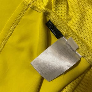Puede incluir: Primer plano de una camiseta deportiva amarilla con un acento verde. La camiseta tiene una textura de malla en el hombro y una etiqueta con el n&uacute;mero 6-52830. La tela es de color amarillo brillante.