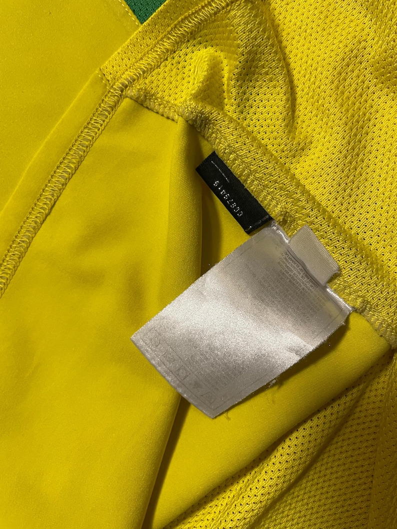 Puede incluir: Primer plano de una camiseta deportiva amarilla con un acento verde. La camiseta tiene una textura de malla en el hombro y una etiqueta con el n&uacute;mero 6-52830. La tela es de color amarillo brillante.