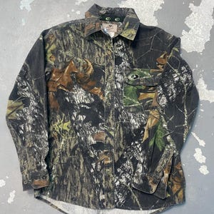 Op de afbeelding: Een camouflage shirt met lange mouwen, een knoopsluiting en een borstzak. Het shirt heeft een patroon van bruin, groen en zwart, met het merklogo zichtbaar op de kraag en de zak.