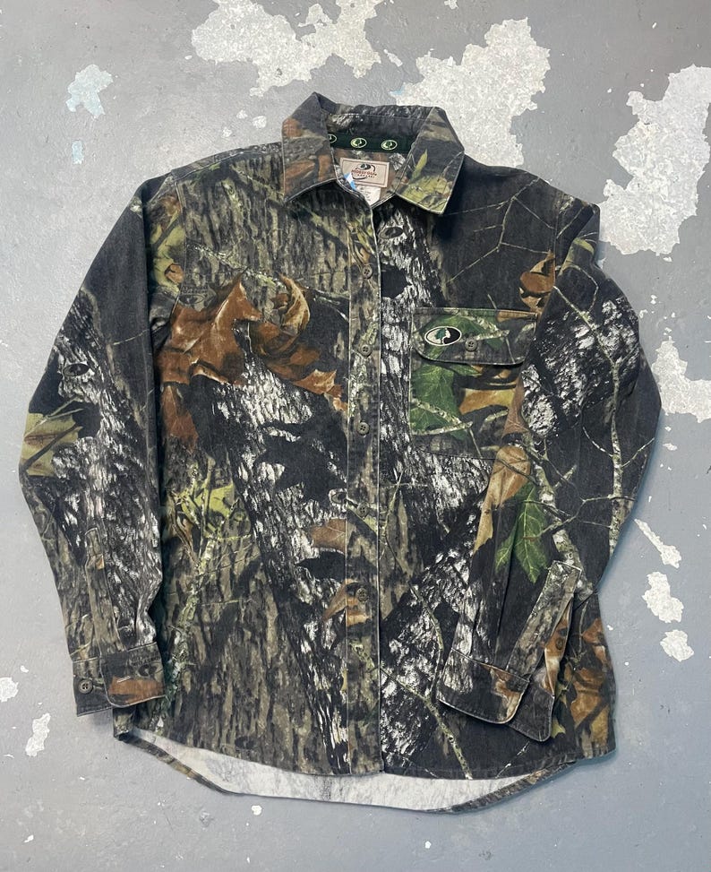 Op de afbeelding: Een camouflage shirt met lange mouwen, een knoopsluiting en een borstzak. Het shirt heeft een patroon van bruin, groen en zwart, met het merklogo zichtbaar op de kraag en de zak.