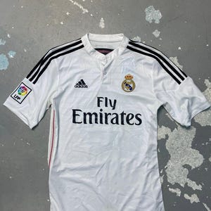 May include: A white short-sleeved soccer jersey featuring black Adidas stripes on the shoulders, a button-up collar, and the text Fly Emirates in black. The Real Madrid crest is on the chest, and an LFP league patch is on the right sleeve.