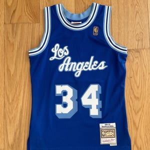 Puede incluir: Una camiseta de baloncesto azul de Los Angeles Lakers con detalles blancos y celeste. La camiseta presenta el número 34 en blanco y celeste, y el nombre "Los Angeles". Se ve una etiqueta Hardwood Classics.