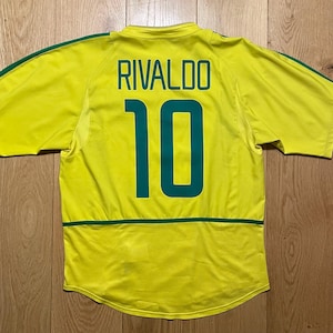 Puede incluir: Una camiseta de f&uacute;tbol amarilla con detalles verdes, con el nombre "RIVALDO" y el n&uacute;mero "10" en verde en la espalda. La camiseta tiene rayas verdes en las mangas y una franja verde en la parte inferior de la espalda.