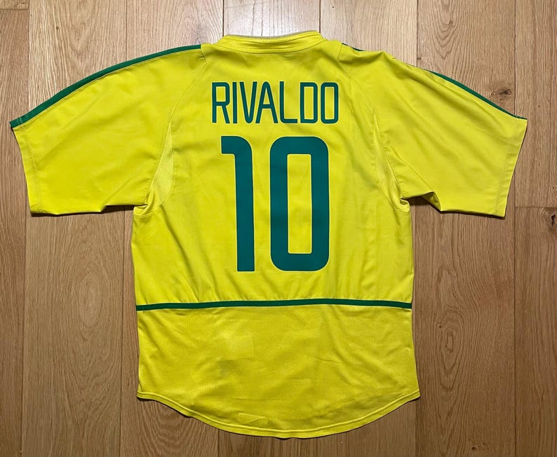 Puede incluir: Una camiseta de f&uacute;tbol amarilla con detalles verdes, con el nombre "RIVALDO" y el n&uacute;mero "10" en verde en la espalda. La camiseta tiene rayas verdes en las mangas y una franja verde en la parte inferior de la espalda.