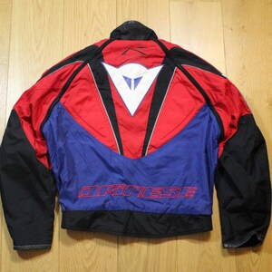 May include: A motorcycle jacket with a red, blue, and black color scheme. The jacket features a white logo on the back and the word "DAINESE" in red lettering. The jacket has black sleeves and a black collar.