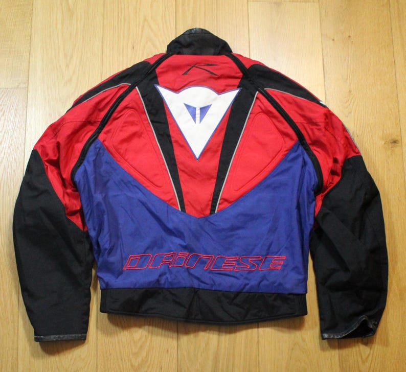 May include: A motorcycle jacket with a red, blue, and black color scheme. The jacket features a white logo on the back and the word "DAINESE" in red lettering. The jacket has black sleeves and a black collar.
