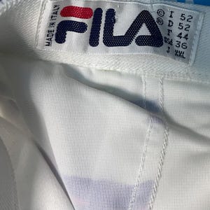 May include: Close-up of a white garment with a colorful trim of pink, purple, and blue. The garment has a tag that reads "FILA" in navy blue and red, with "MADE IN ITALY" above it. The tag also includes size information.