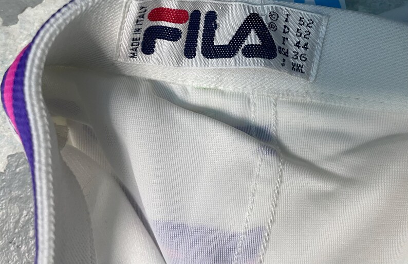 May include: Close-up of a white garment with a colorful trim of pink, purple, and blue. The garment has a tag that reads "FILA" in navy blue and red, with "MADE IN ITALY" above it. The tag also includes size information.