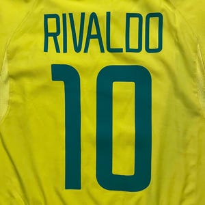 Puede incluir: Una camiseta de f&uacute;tbol amarilla con el nombre "RIVALDO" y el n&uacute;mero "10" impresos en verde azulado. La camiseta tiene un ribete verde en la parte inferior y alrededor del cuello.