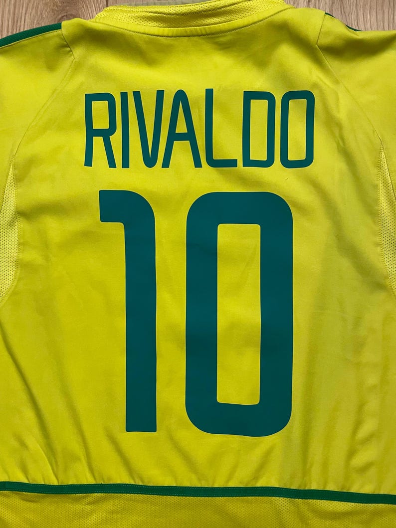 Puede incluir: Una camiseta de f&uacute;tbol amarilla con el nombre "RIVALDO" y el n&uacute;mero "10" impresos en verde azulado. La camiseta tiene un ribete verde en la parte inferior y alrededor del cuello.