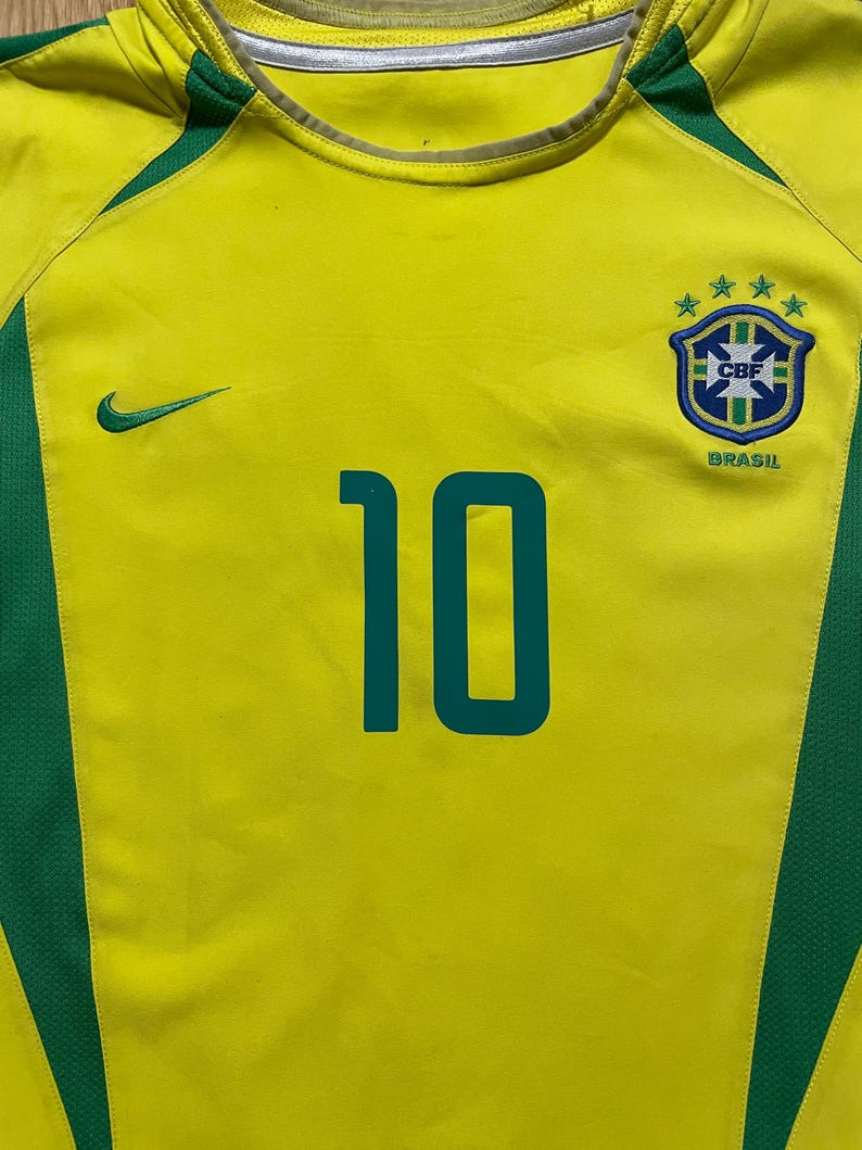 Puede incluir: Camiseta de f&uacute;tbol de Brasil amarilla y verde con el n&uacute;mero 10 en verde azulado. Presenta el logo de Nike y el escudo de la selecci&oacute;n brasile&ntilde;a. La camiseta tiene detalles verdes en hombros y laterales.