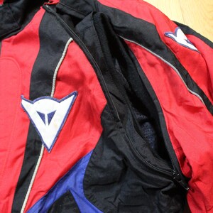 May include: A red, black, and blue motorcycle jacket with white accents. The jacket features a zipper closure, a stand-up collar, and a white logo patch on the chest. The jacket is made of a lightweight, possibly nylon, material.