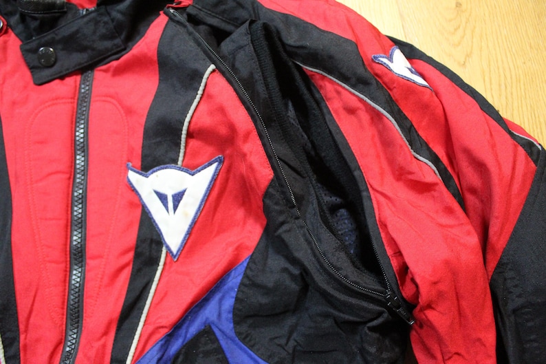 May include: A red, black, and blue motorcycle jacket with white accents. The jacket features a zipper closure, a stand-up collar, and a white logo patch on the chest. The jacket is made of a lightweight, possibly nylon, material.