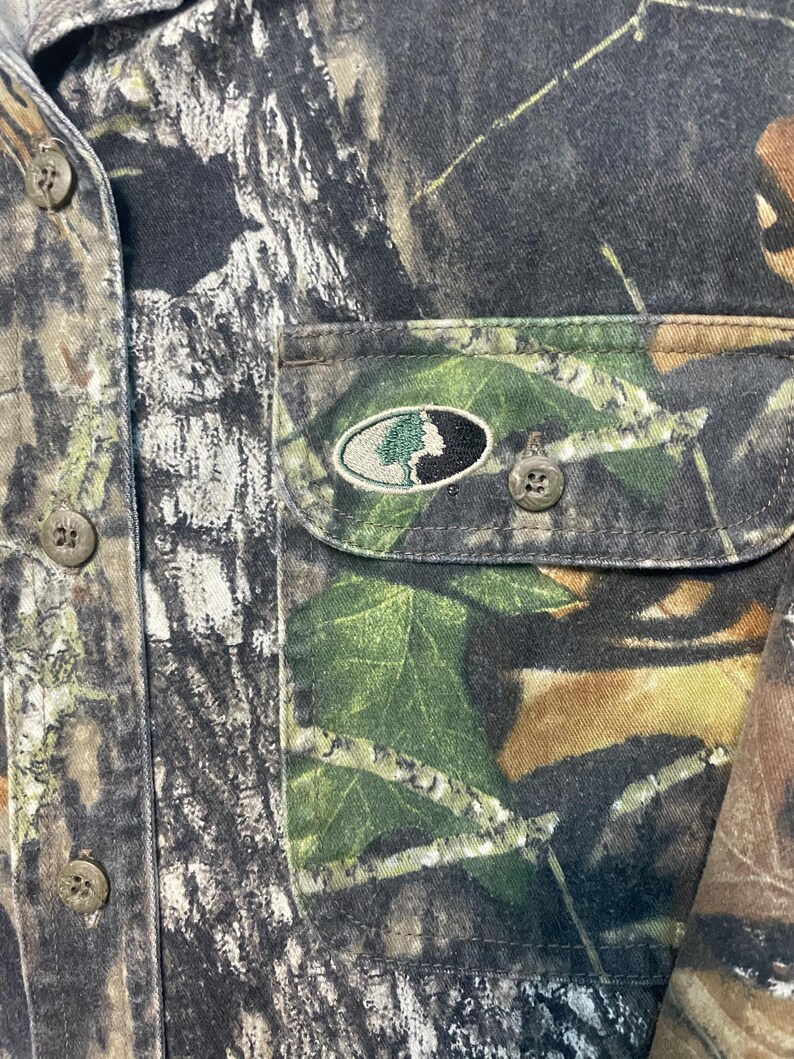 Op de afbeelding: Close-up van een camouflage overhemd met knopen. Het overhemd heeft een bos camouflagepatroon in tinten bruin, groen en grijs. Een borstzak heeft een logo met een boomontwerp. Het overhemd heeft verschillende bruine knopen.