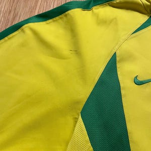 Puede incluir: Primer plano de una camiseta deportiva amarilla y verde. La camiseta tiene un cuerpo amarillo con detalles verdes en los hombros y los lados. Un logotipo verde de Nike es visible en el lado derecho.