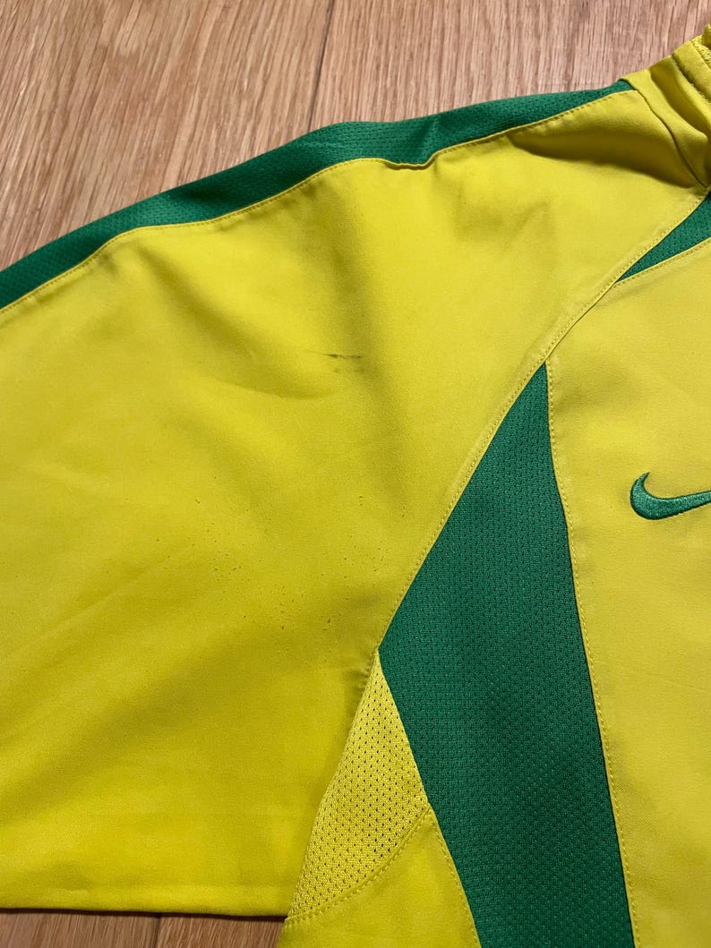 Puede incluir: Primer plano de una camiseta deportiva amarilla y verde. La camiseta tiene un cuerpo amarillo con detalles verdes en los hombros y los lados. Un logotipo verde de Nike es visible en el lado derecho.