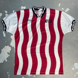 Pode incluir: Uma camisola de futebol de manga curta com um padr&atilde;o de riscas verticais onduladas em vermelho e branco. A camisola tem um decote em V azul-escuro com rebordo branco, o log&oacute;tipo Adidas preto &agrave; direita e um emblema US azul-escuro &agrave; esquerda.