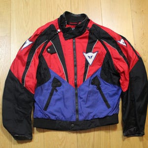 May include: A red, black, and blue motorcycle jacket with a zip-up front and a stand-up collar. The jacket features white logos and black accents. The jacket has a black ribbed hem and cuffs. The jacket is made of a lightweight material.