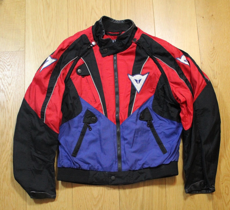 May include: A red, black, and blue motorcycle jacket with a zip-up front and a stand-up collar. The jacket features white logos and black accents. The jacket has a black ribbed hem and cuffs. The jacket is made of a lightweight material.