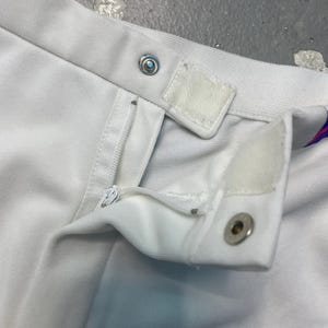 May include: Close-up of white garment with a zipper, snap closure, and Velcro tab. The fabric appears to be a soft, possibly cotton or a blend. The garment is likely a skirt or pants.