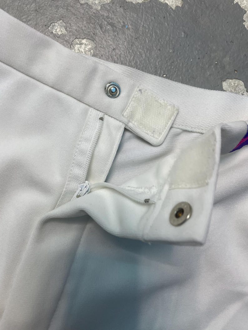 May include: Close-up of white garment with a zipper, snap closure, and Velcro tab. The fabric appears to be a soft, possibly cotton or a blend. The garment is likely a skirt or pants.