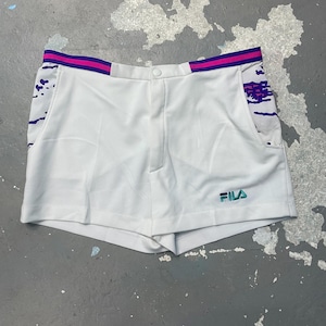 May include: White athletic shorts with a button closure and a small pocket. The shorts have a purple and pink striped waistband and accents. The Fila logo is embroidered in teal on the lower right side.