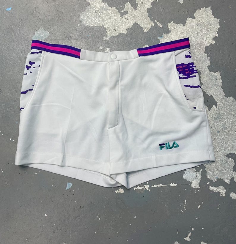 May include: White athletic shorts with a button closure and a small pocket. The shorts have a purple and pink striped waistband and accents. The Fila logo is embroidered in teal on the lower right side.