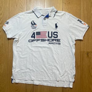 May include: A white short-sleeved polo shirt featuring a navy blue collar trim and a button placket. The chest displays a navy blue graphic with the text 4 US OFFSHORE RACING, a small American flag, a sailboat logo, and a dark blue polo player emblem.