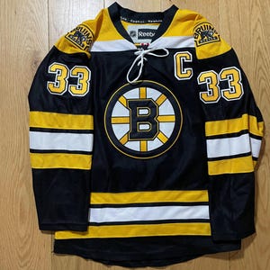Puede incluir: Camiseta de hockey de los Boston Bruins en negro y dorado con el número 33 en las mangas y un gran logotipo de los Bruins en el pecho. La camiseta tiene rayas blancas y doradas y la palabra "Reebok" en el cuello.