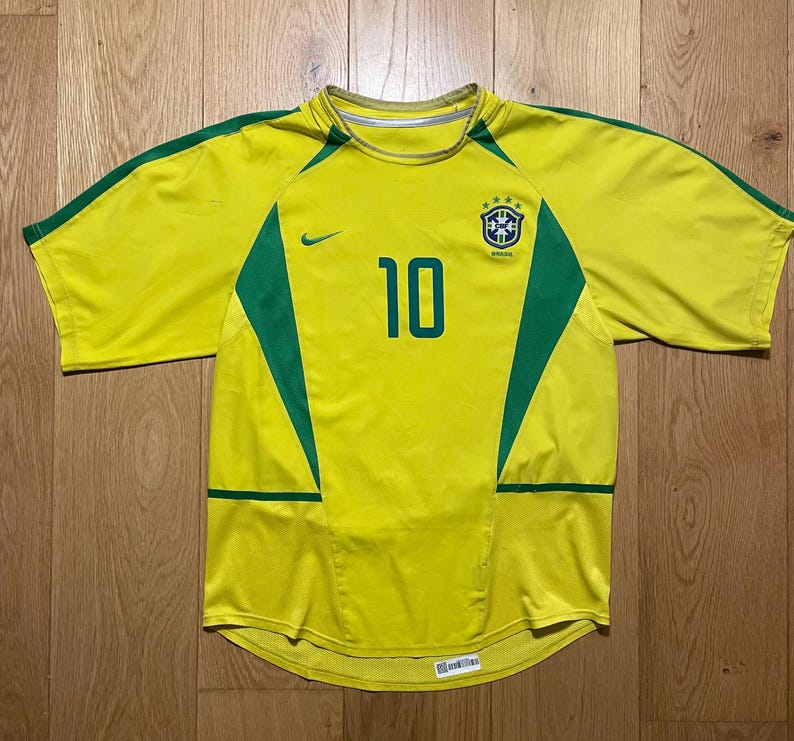 Puede incluir: Una camiseta de f&uacute;tbol amarilla de Brasil con detalles verdes y el n&uacute;mero 10 en el centro. La camiseta presenta un logotipo de Nike y el escudo del equipo de Brasil. La camiseta es de manga corta.