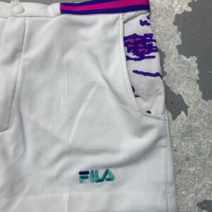 May include: White athletic shorts with a colorful design on the pocket and waistband. The shorts feature the brand name "FILA" embroidered in teal and purple. The waistband has pink and purple stripes.