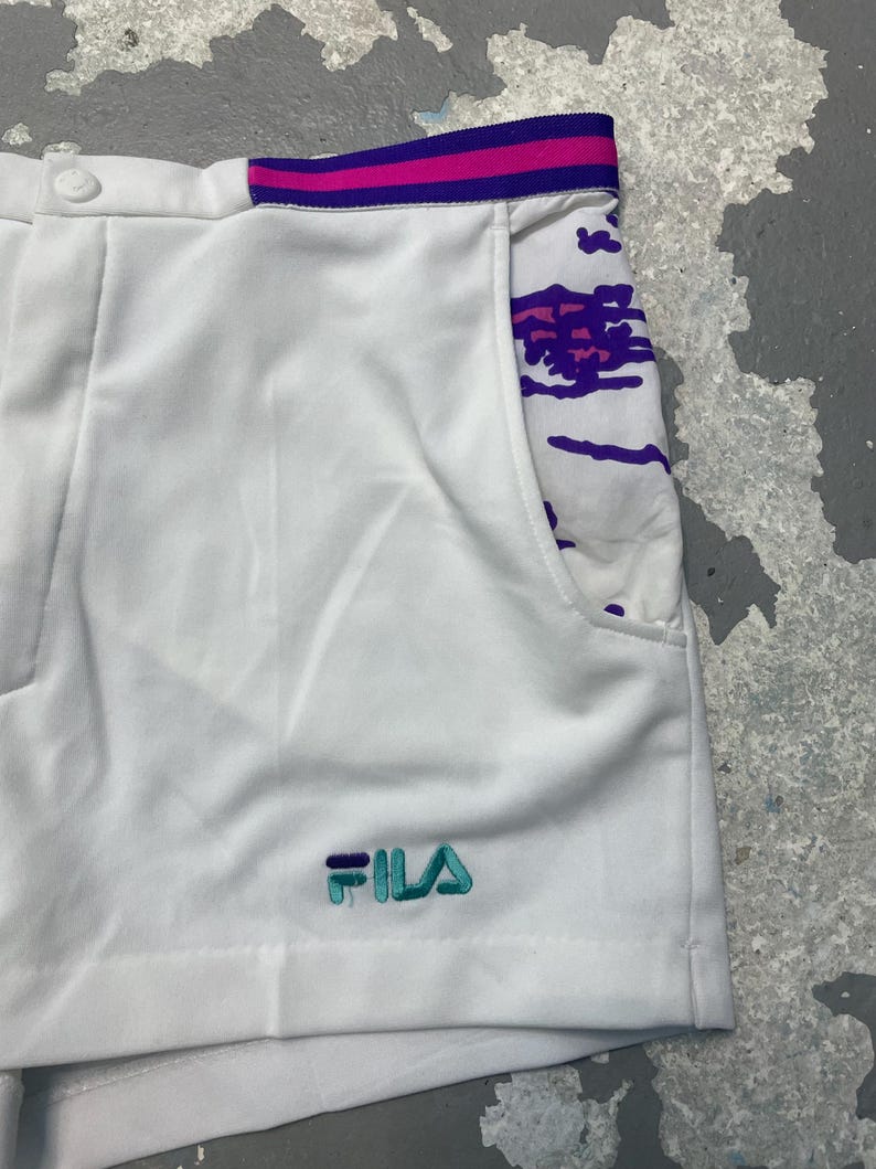 May include: White athletic shorts with a colorful design on the pocket and waistband. The shorts feature the brand name "FILA" embroidered in teal and purple. The waistband has pink and purple stripes.