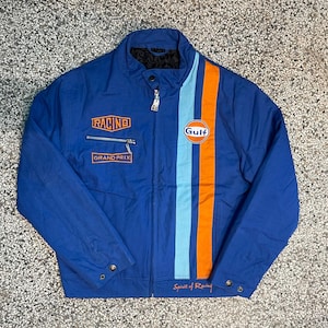 Puede incluir: Chaqueta de carreras azul con rayas naranjas y celeste. Presenta el logo "Gulf" y el texto "Racing" y "Grand Prix". Incluye cierre de cremallera y botones a presión.