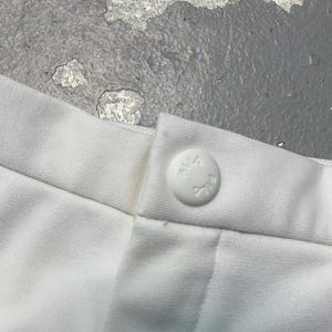 May include: Close-up of white fabric with a button closure. The button is round and white, with embossed text. The fabric appears to be a soft, woven material. The image is well-lit, showing the details of the fabric and button.
