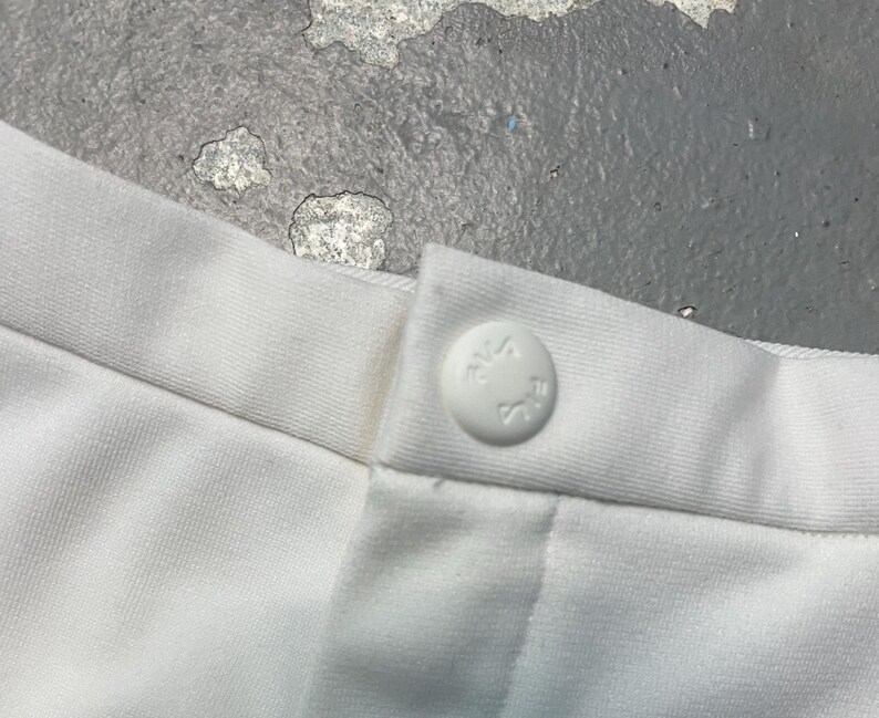 May include: Close-up of white fabric with a button closure. The button is round and white, with embossed text. The fabric appears to be a soft, woven material. The image is well-lit, showing the details of the fabric and button.