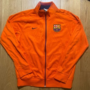 Könnte beinhalten: Orangefarbene Trainingsjacke mit Reißverschluss, mit einem marineblauen Nike-Swoosh und einem Vereinswappen. Die Jacke hat einen hohen Kragen und einen durchgehenden Reißverschluss. Bündchen und Saum sind gerippt. Sportbekleidung.