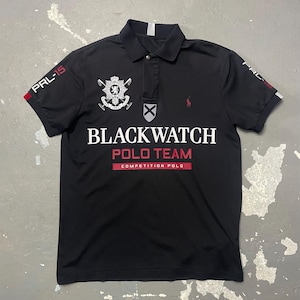 May include: Black polo shirt with short sleeves. The shirt features the text "BLACKWATCH POLO TEAM" in white and red lettering. The shirt also has a crest and the text "PRL-15" on the sleeves. A small red logo is visible on the chest.