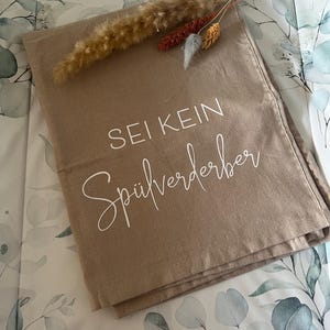May include: A tan-colored rectangular textile with the German text "SEI KEIN Spülverderber" in white script. The textile is folded and adorned with dried ornamental grasses and flowers. The background features a floral pattern.