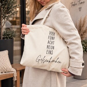 May include: A cream-colored canvas tote bag featuring black printed text. The bag displays the words VIER, FÜNF, ACHT, NEUN, EINS stacked vertically in a sans-serif font, with the word Gelsenkirchen written below in a cursive script.