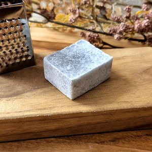 Könnte beinhalten: Eine kleine silberne Reibe und eine graue Seifenstange auf einem Holzbrett. Das Brett liegt auf einem größeren Holzbrett.