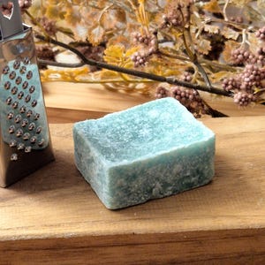 Könnte beinhalten: Eine kleine, silberne Reibe mit herzförmigen Löchern liegt neben einer hellblauen Seifenstange auf einem Holzbrett.