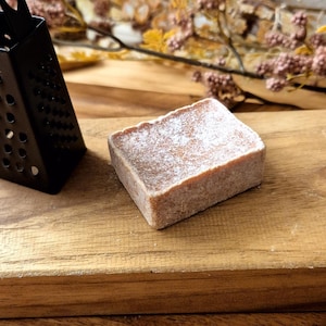 Könnte beinhalten: Ein kleiner, rechteckiger Stück Seife mit hellbrauner Farbe und körniger Textur liegt auf einem Holzbrett. Eine schwarze Metallreibe mit einem herzförmigen Griff befindet sich ebenfalls auf dem Brett.